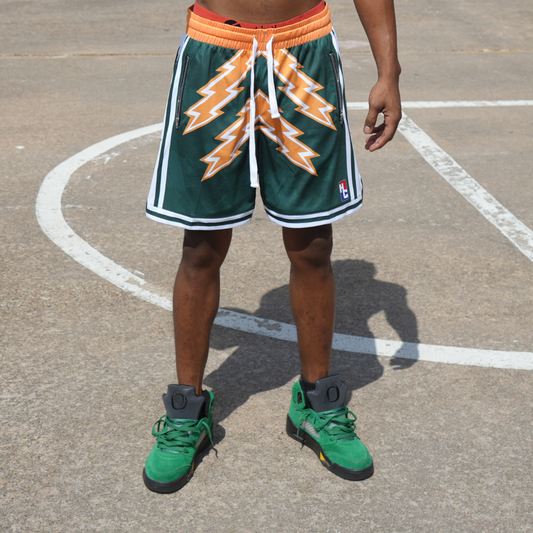 Man wearing Emerald City Shorts in green with orange lightning design and white drawstrings on outdoor basketball court.